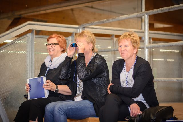 Das Bild zeigt drei Frauen, nebeneinander auf einer Bank sitzend. Die Frau in der Mitte schaut durch ein Fernglas. 