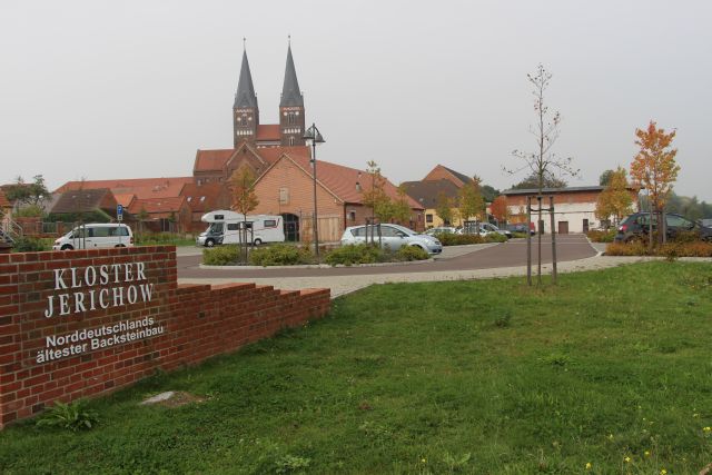 Backsteingebäude mit den beiden Klostertürmen dahinter, davor ein Parkplaötz mit Wendeschleife.