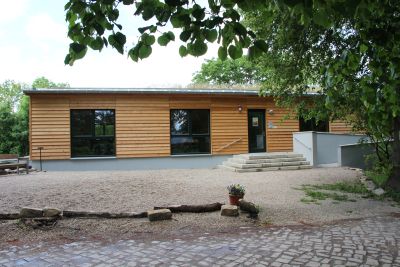 Vorderansicht des Schulungsgebäudes. Gebaut im Bungalowstil mit Holzverkleidung. Große Fenster, zum Eingang führen eine Treppe und eine Rollstuhlfaherrampe. 