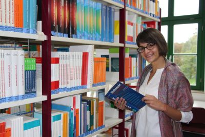 Das Bild zeigt eine junge Frau vor einem Bücherregal.