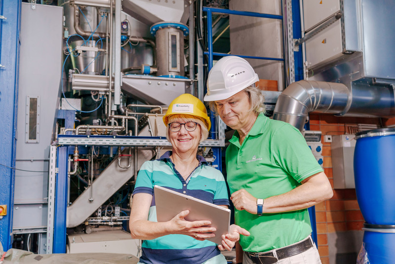 Das Foto zweigt zwei Personen mit Tablett vor einer Industrieanlage. 