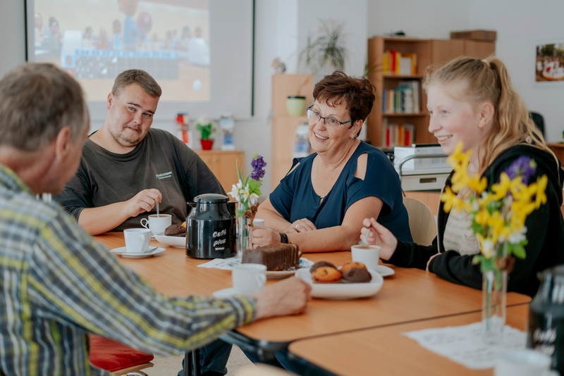 Das Bild zeigt vier Personen an einem Tisch mit Kaffee und Keksen. 