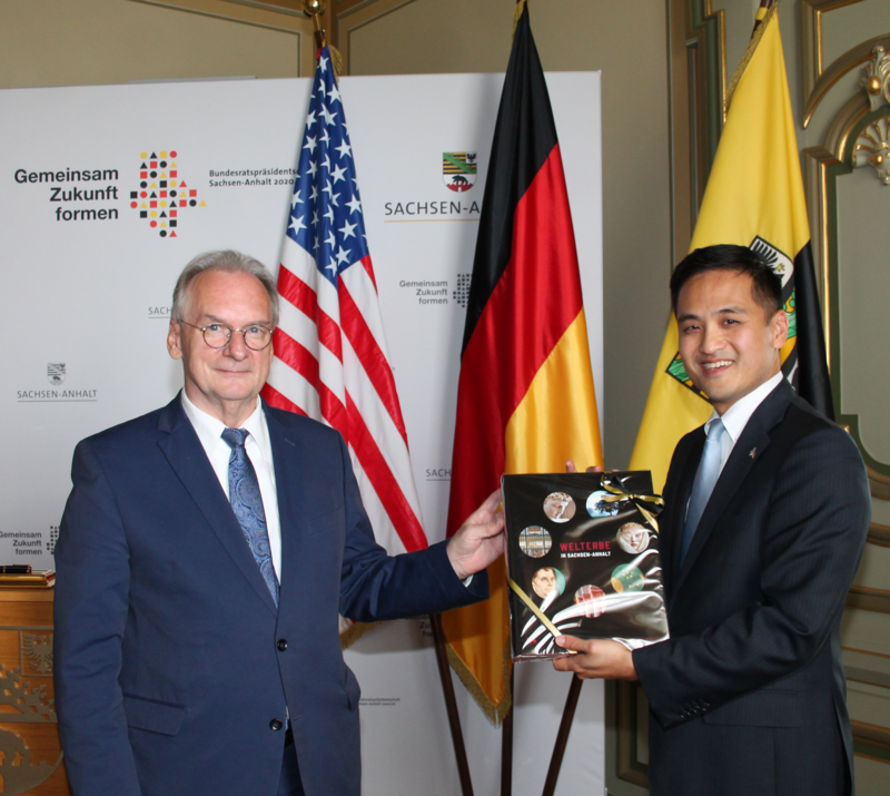 Das Bild zeigt Ministerpräsident Dr. Reiner Haseloff (links) bei der Überreichung eines Bildbandes zum Welterbe Sachsen-Anhalt an den US-Generalkonsul (rechts) Ken Toko. Beide stehen vor den Fahnen der USA, Deutschland und Sachsen-Anhalt.